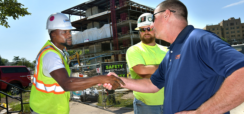 Men in hardhats and yellow vests shaking hands in front of a building under construction