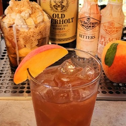 A cocktail glass with a peach wedge in front of various bottles of alcohol